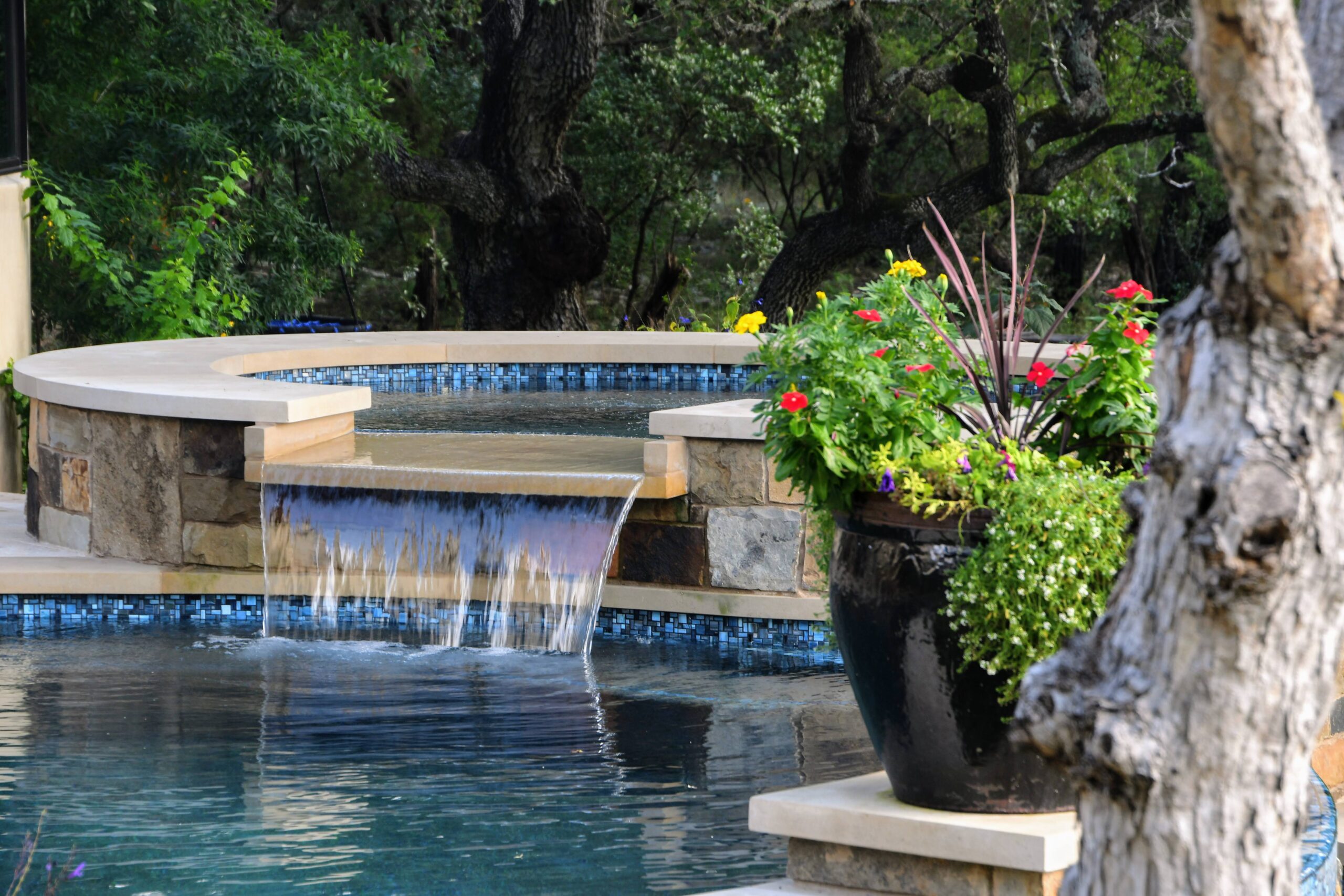 A serene pool scene featuring a small cascading waterfall connecting a raised hot tub to the main pool. The area is framed by lush greenery and a large potted plant with vibrant red and yellow flowers.