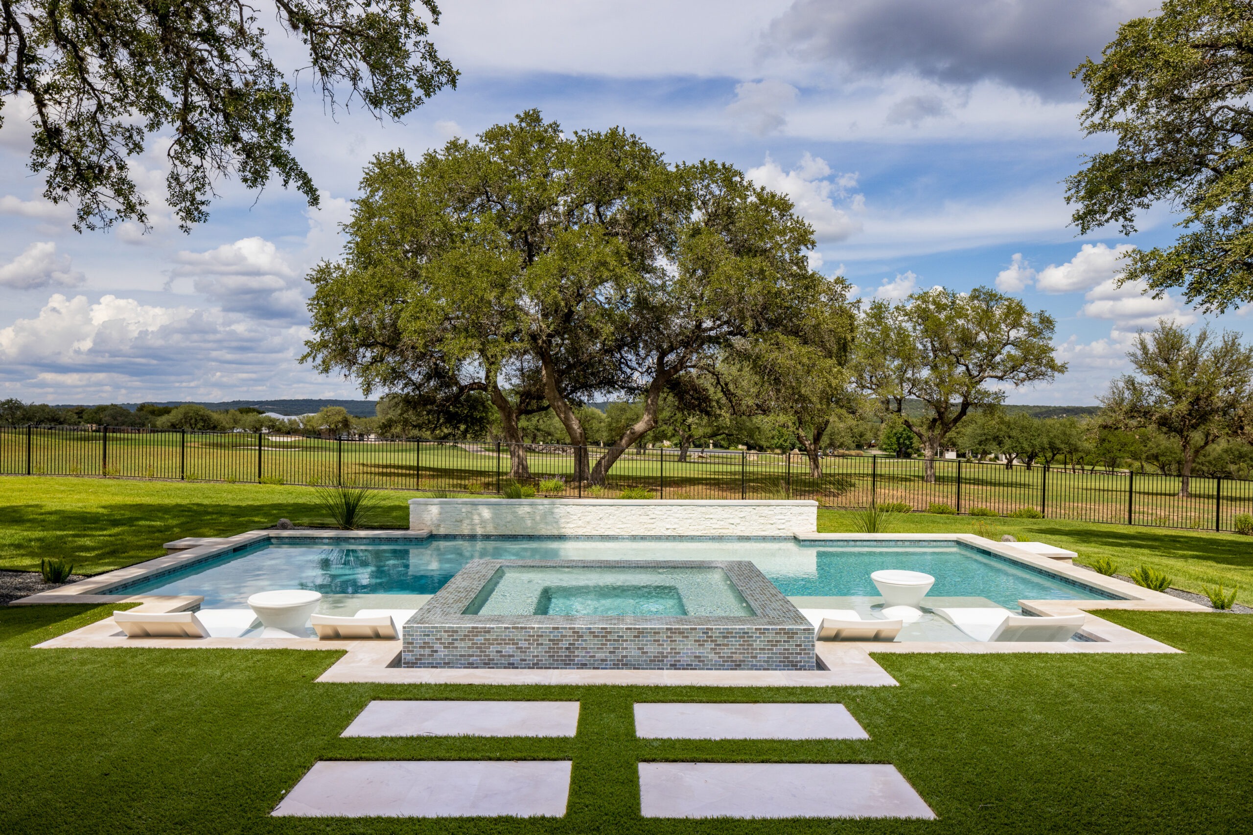 A luxurious backyard features a rectangular swimming pool with a central hot tub. Four white chairs border the pool, surrounded by lush grass and large trees. The sky is partly cloudy, and fields extend into the distance beyond a black metal fence.