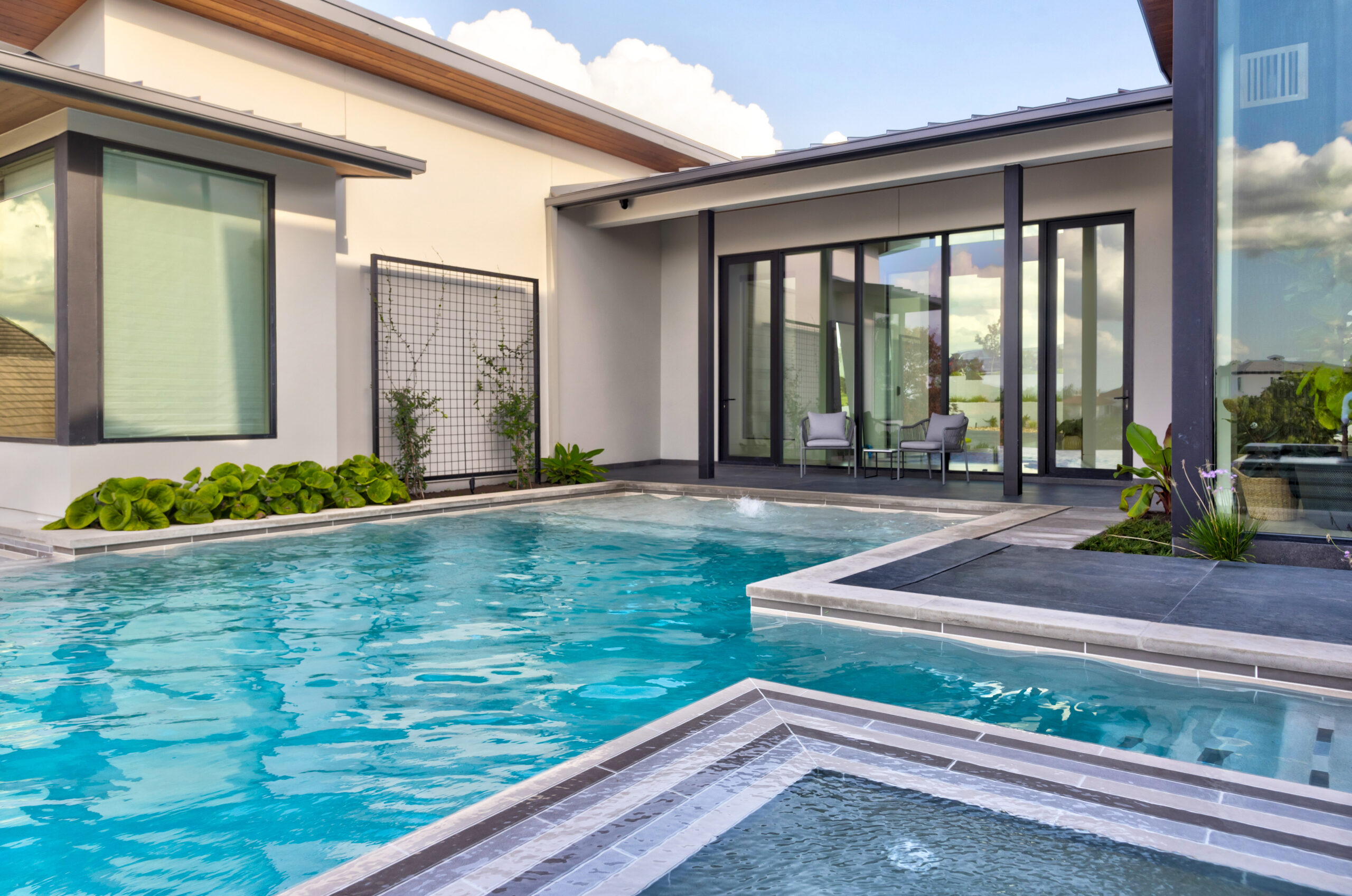 Modern backyard with a swimming pool and spa. The house features large windows and a covered patio with seating. Lush greenery decorates the area, and the sky is partly cloudy.