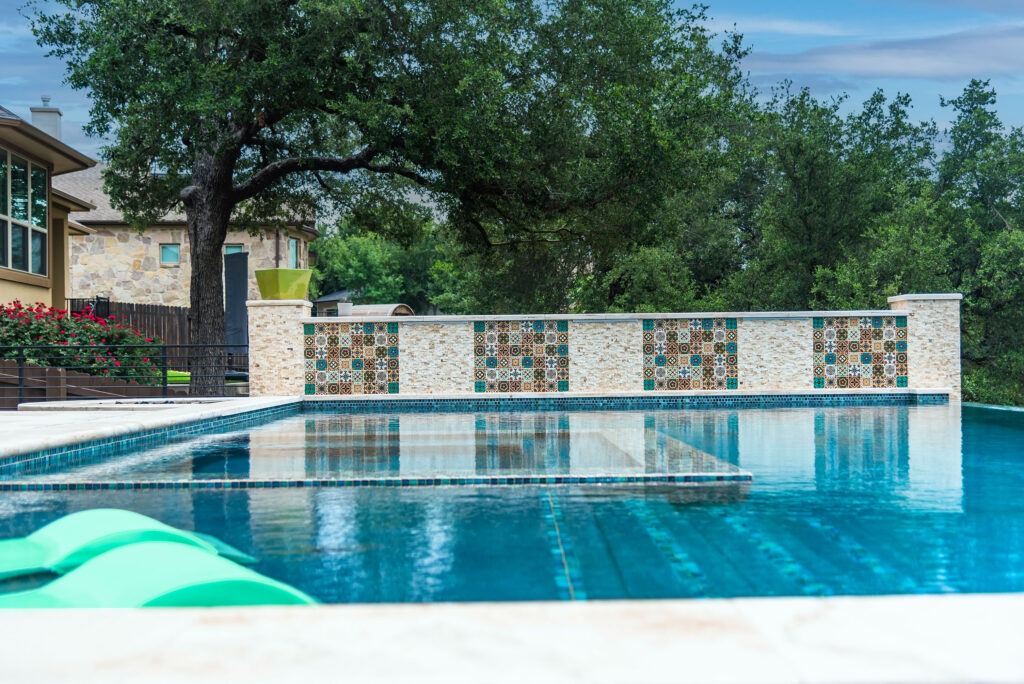 A tranquil swimming pool with turquoise water is surrounded by decorative tile walls. Trees and a house are visible in the background, and a green inflatable pool float rests on the water's surface. The sky is partly cloudy.