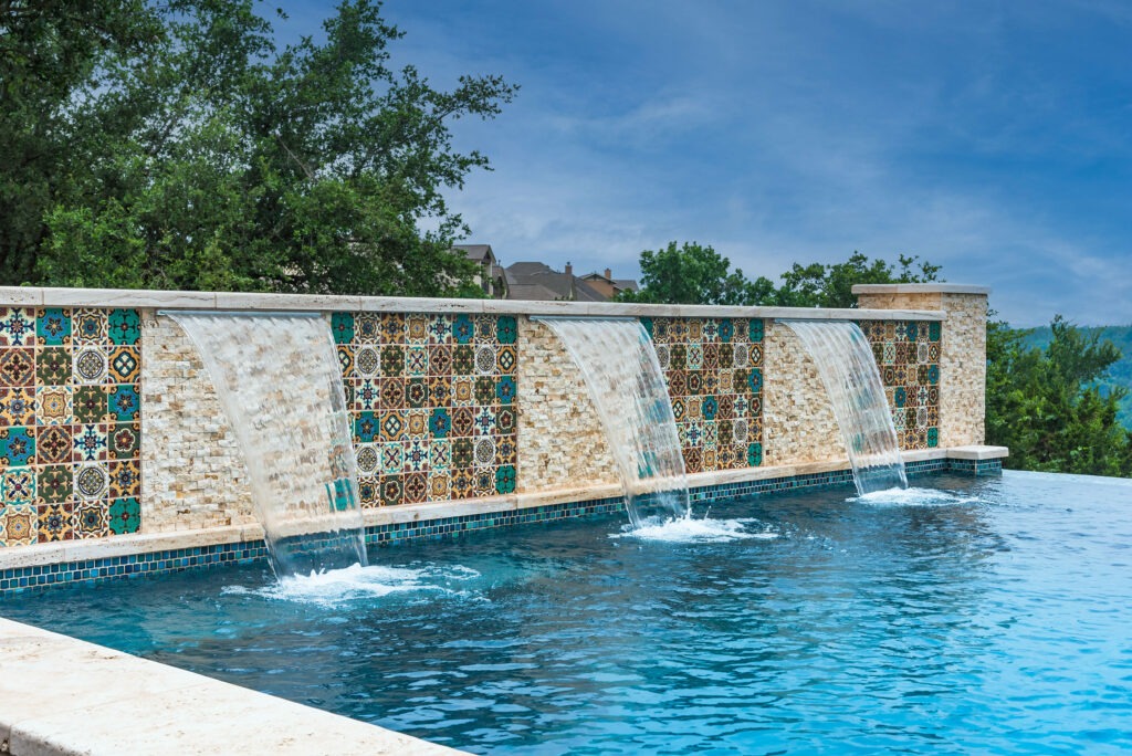 Swimming pool with a tiled stone water feature, where water cascades over the edge into the pool. The tiles have colorful geometric patterns. Trees and a clear sky are visible in the background.