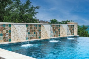 Swimming pool with a tiled stone water feature, where water cascades over the edge into the pool. The tiles have colorful geometric patterns. Trees and a clear sky are visible in the background.