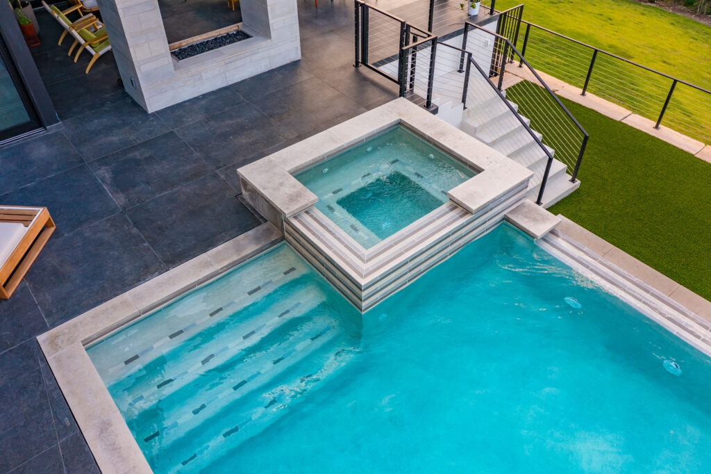 A modern rectangular swimming pool with a connected square hot tub. The pool area has a sleek gray stone patio, glass railings, and is adjacent to a grassy lawn, leading up to a contemporary building with outdoor seating.