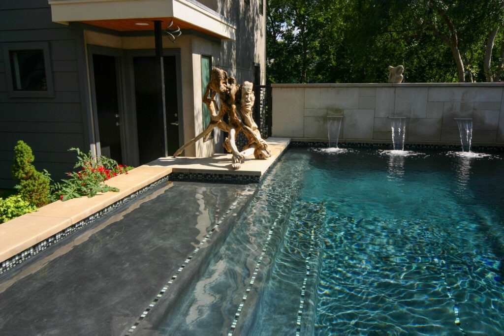 Modern backyard pool with a sleek design, featuring a shallow entry area. A large, artistic bronze sculpture stands at the edge near the house. Water flows from three spouts on a stone wall, and trees are visible in the background.