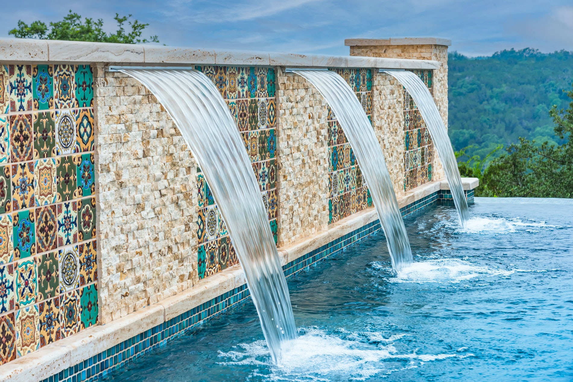 Water cascades from three stone and tile fountains into a pool, set against a backdrop of lush, green hills and a cloudy sky. The tiled wall features intricate, colorful patterns that add a decorative touch to the scene.