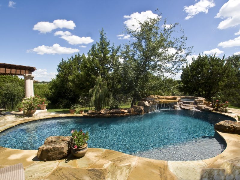 A serene backyard pool surrounded by lush greenery and rocks. The pool features a small waterfall and appears inviting under a clear blue sky with scattered clouds. There are potted plants and a pergola in the background, enhancing the tranquil setting.