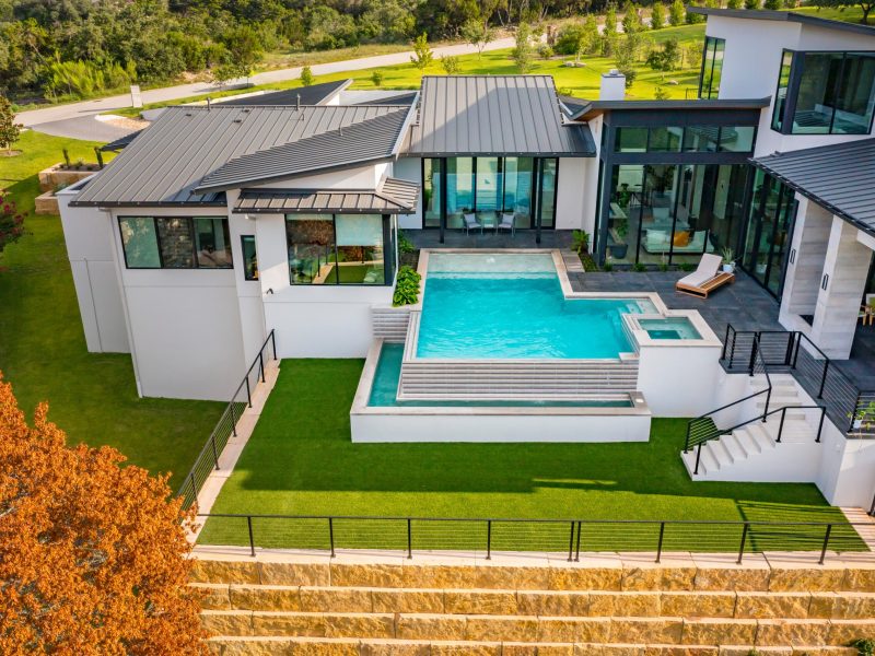 Aerial view of a modern white house with a grey metal roof, a swimming pool, and a hot tub. The house features expansive windows, a patio with a lounge chair, and a landscaped lawn. It's situated near greenery and a road.