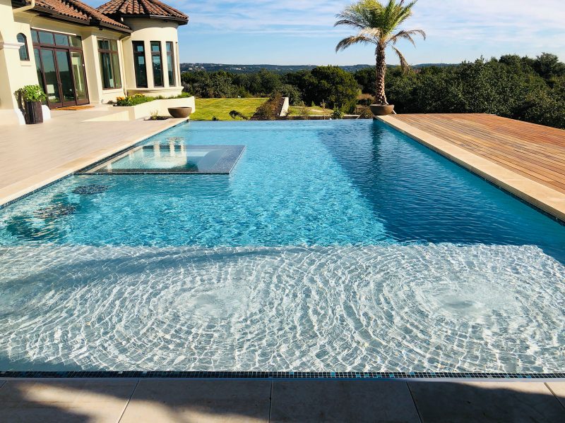 A luxurious infinity pool with a wooden deck and a palm tree overlooks a scenic landscape. The pool has a built-in lounging area with clear water and gentle ripples. A modern house with large windows is on the left.