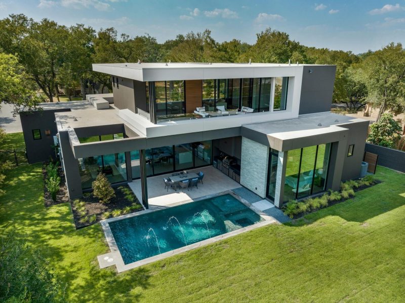 A modern, two-story house with large windows, a flat roof, and an L-shaped design. It features a spacious backyard with a rectangular swimming pool and a surrounding grassy lawn, set against a backdrop of lush trees under a clear blue sky.