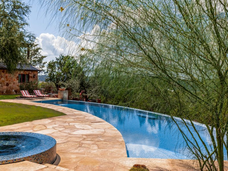 Curved stone pathway leads to an infinity pool surrounded by lush trees and grass. Two striped lounge chairs are near the edge. The sky is partly cloudy, and a stone building is visible in the background.