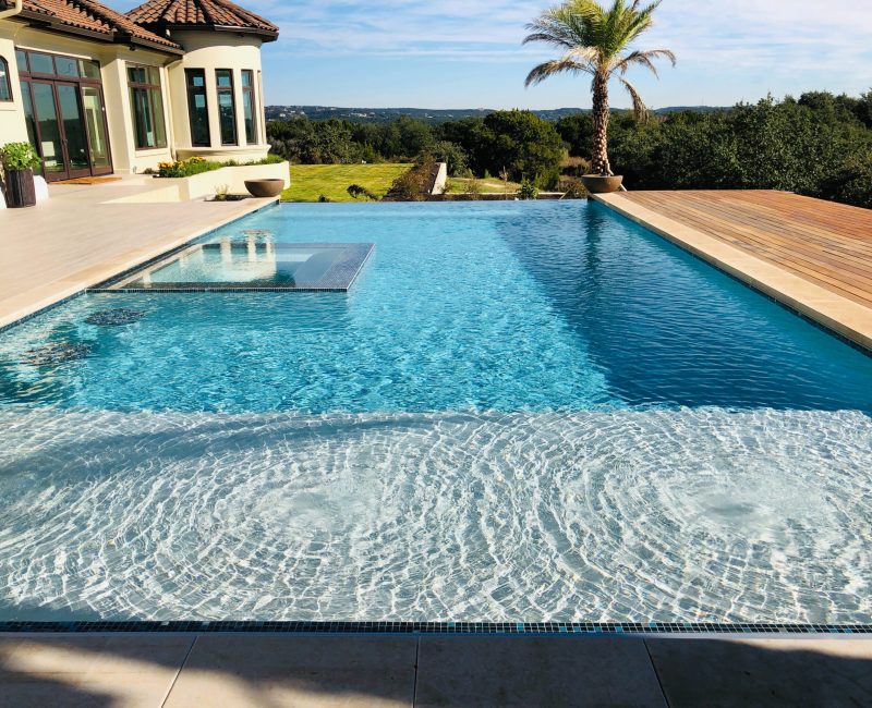 A luxurious infinity pool with clear blue water overlooks a scenic landscape. The pool features a shallow lounging area and an adjacent hot tub. In the background, a large house and a single palm tree are visible under a clear sky.