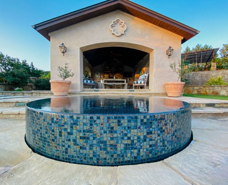 A small, round infinity pool with blue mosaic tiles sits in front of a cozy building with a covered patio. Potted plants flank the entrance, and outdoor furniture is visible inside. The setting is surrounded by greenery under a clear blue sky.