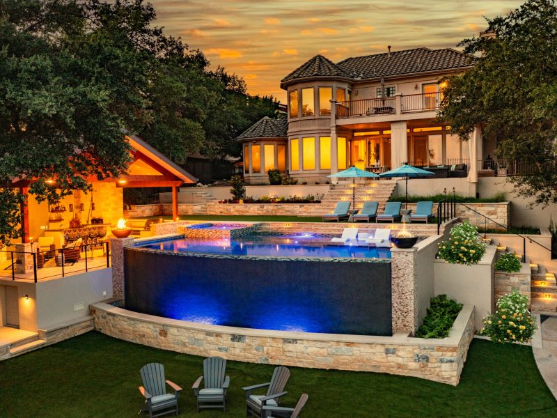 A luxurious two-story house with large windows is set against a vibrant sunset. An illuminated infinity pool and jacuzzi with blue lights are in the foreground. A wooden patio and seating area with Adirondack chairs are surrounded by lush greenery.