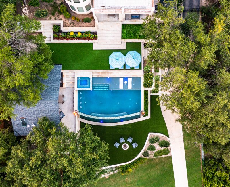 Aerial view of a modern backyard with a rectangular pool, a small spa, and patio furniture. The area is surrounded by green grass and lush trees. A nearby building has a grey roof, and a circular seating area is visible on the lawn.