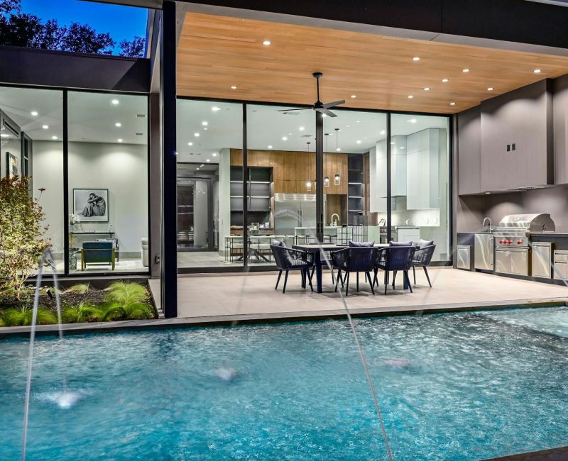 Modern house with a glass-walled interior visible from the outside, featuring a kitchen and dining area. The foreground has a pool with water fountains. A patio with dining furniture and an outdoor grill is also present.