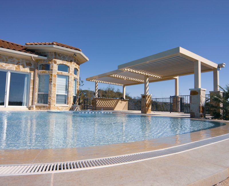 A luxurious house with large windows sits beside a clear, round pool. A pergola extends over a patio area, offering shade. The sky is clear and blue, with some greenery visible in the background.