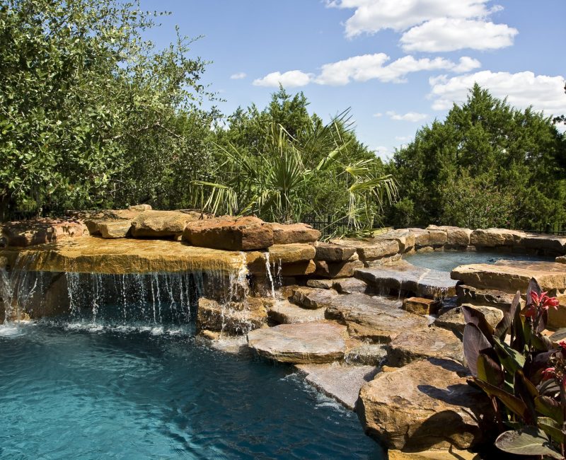 A serene pool with cascading water flows over natural stone ledges, surrounded by lush greenery and tropical plants under a bright blue sky with fluffy clouds.