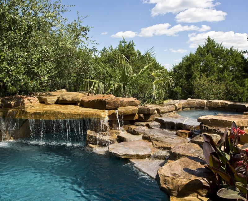 Copy of dinnes day-3 A serene pool with cascading water flows over natural stone ledges, surrounded by lush greenery and tropical plants under a bright blue sky with fluffy clouds.