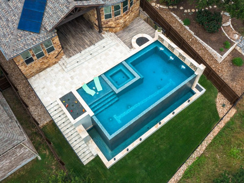 Aerial view of a modern backyard with a rectangular pool featuring a raised spa. The pool is bordered by stone tiles and surrounded by a lawn. A wooden deck with a house entrance is adjacent to the pool. Decorative plants line the pathway.