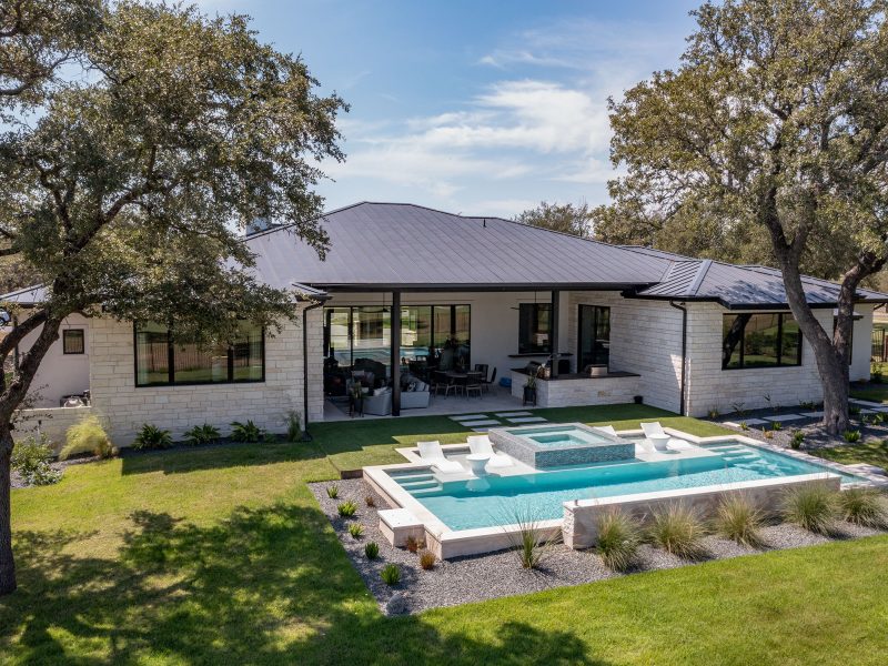 Modern one-story house with a large patio and swimming pool surrounded by a landscaped yard. The house has a grey roof, light stone exterior, and is shaded by large trees under a clear blue sky.