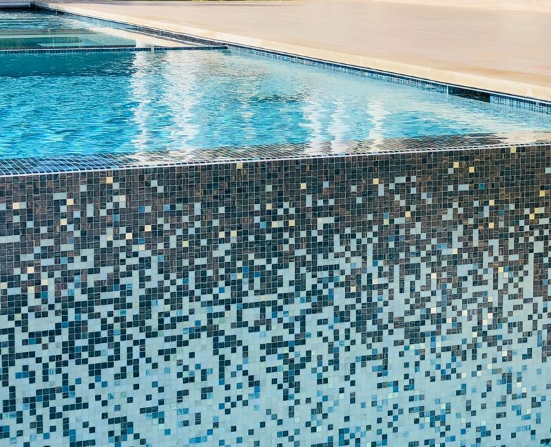 A close-up view of a swimming pool with mosaic tile detailing. The multicolored tiles form a gradient pattern on the pool's edge. The water is clear, reflecting light, and the pool deck is visible in the background.