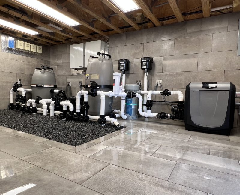 A mechanical room with water filtration equipment. Large filters and pipes are mounted on a tiled wall, connected by black and white fittings. The floor is shiny with gray tiles, and the ceiling has exposed wooden beams.