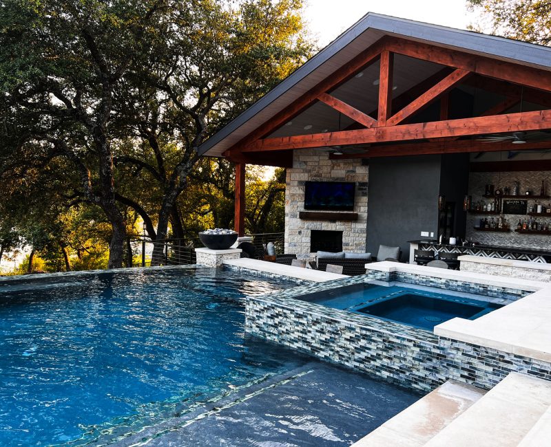 A luxurious outdoor space featuring a modern pool with a built-in hot tub. There's a covered patio area with wood beams, a stone fireplace, bar, and seating. Mature trees surround the area, enhancing the serene ambiance.