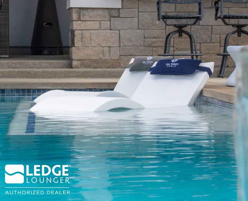 Two white loungers partially submerged at the edge of a swimming pool with clear blue water. The backdrop features a stone wall and bar stools. The "Ledge Lounger" logo and "Authorized Dealer" text appear in the lower left corner.