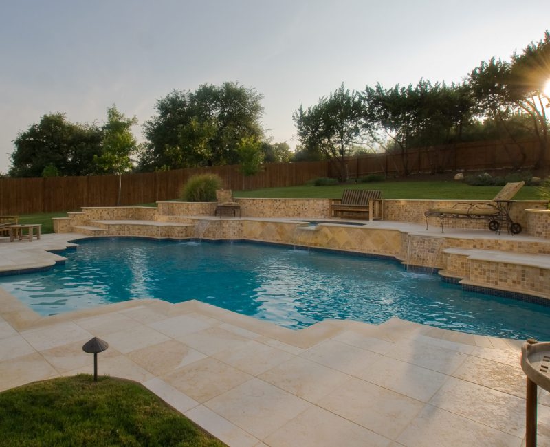 A backyard with a rectangular swimming pool surrounded by stone tiles. There are wooden lounge chairs and a bench nearby. A wooden fence and tall trees line the back, and the sun is setting in the background, casting a warm glow.