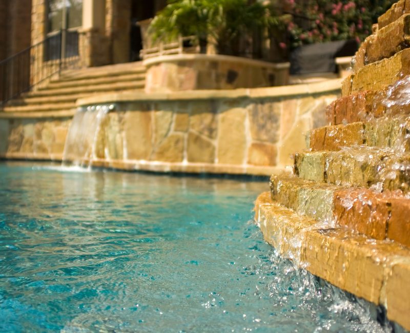 A pool with clear blue water features a cascading stone waterfall in the foreground. Steps lead up to a patio area with lush green plants in the background, suggesting a luxurious outdoor setting.